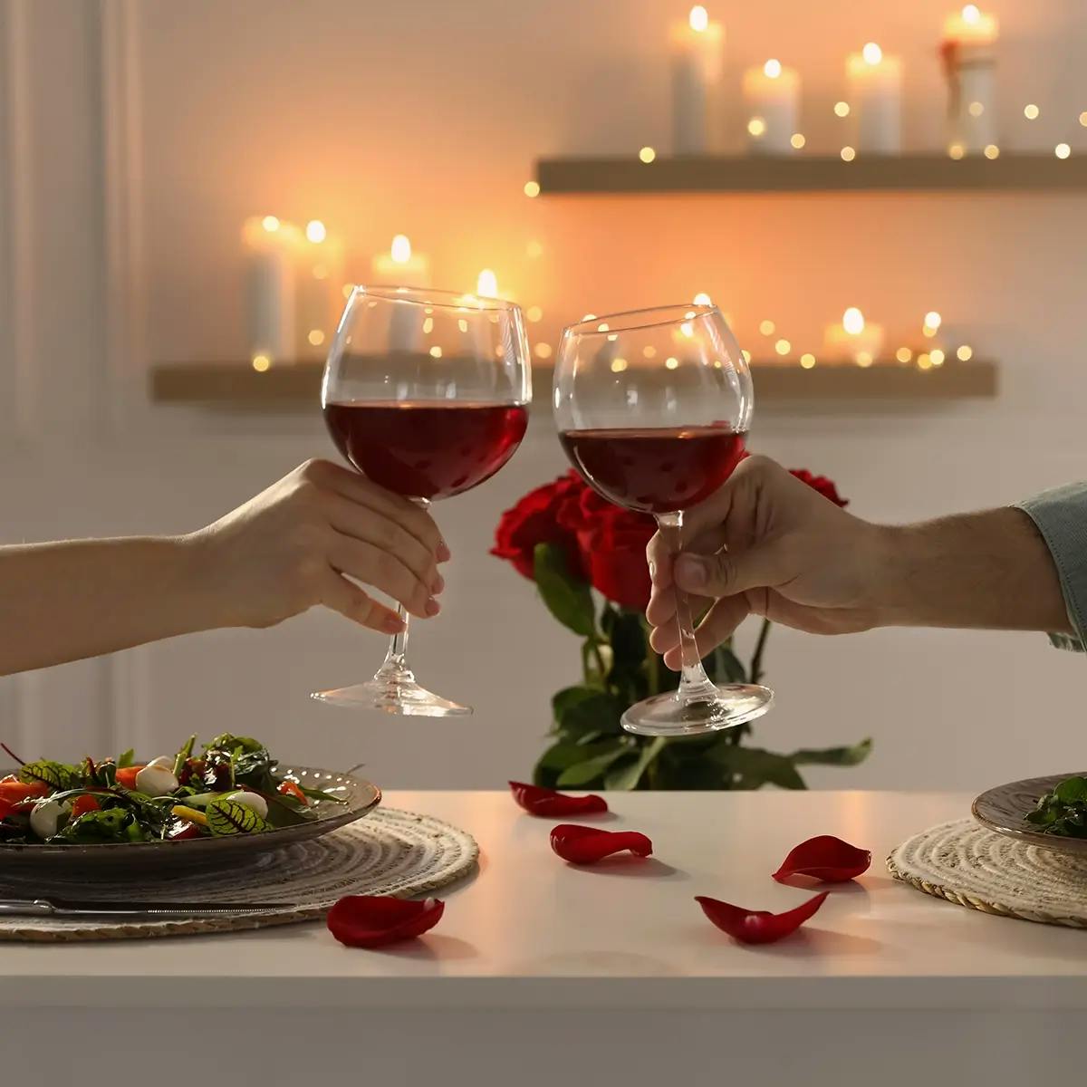 Two hands toasting with red wine glasses at a table set with dinner for two, with candles in the background.