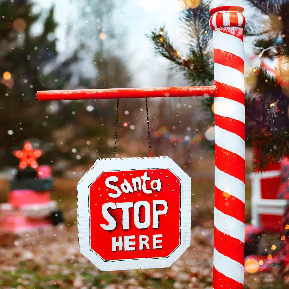 “Santa, Stop Here” sign on a pole in the front yard of a house with snow falling in the background.
