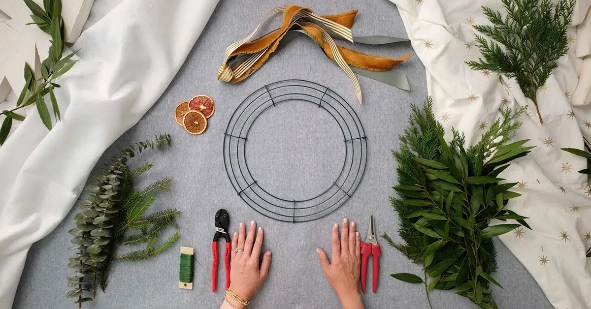 Hands on a table surrounded by supplies to make a Christmas or Thanksgiving wreath.