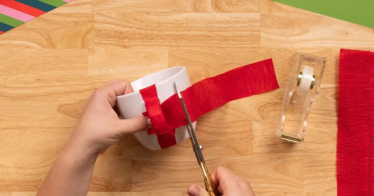 Wrapping a piece of crepe paper between the top and bottom of the handle of a coffee cup, in a tutorial on how to wrap a mug.