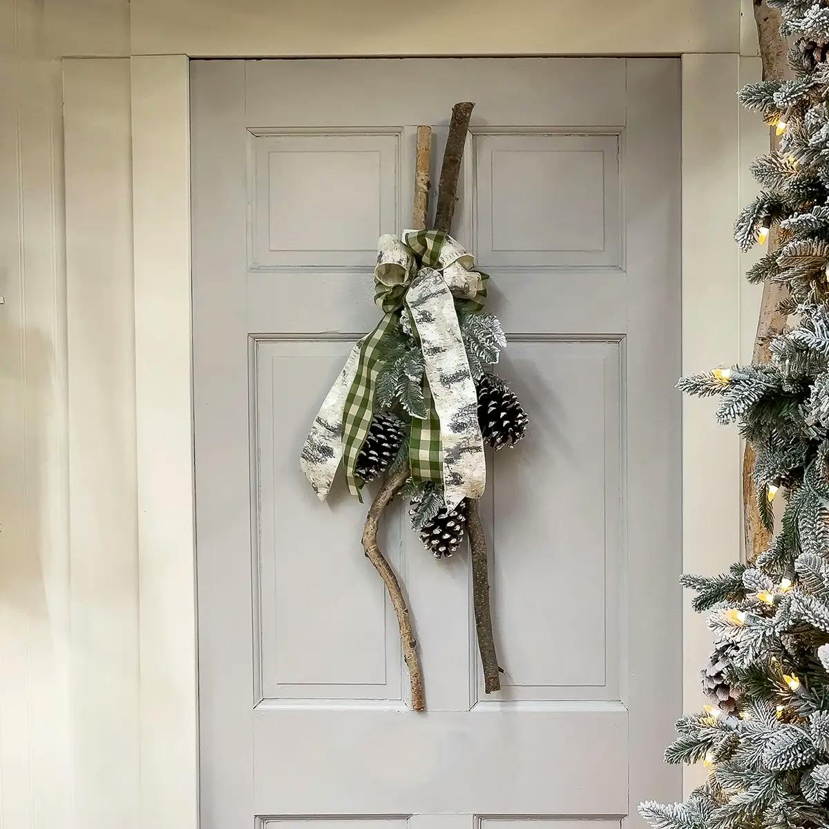 Front porch with white front door with a handmade wreath of branches and pinecones on the door. A frosted Christmas tree in foreground with acorns and bows.