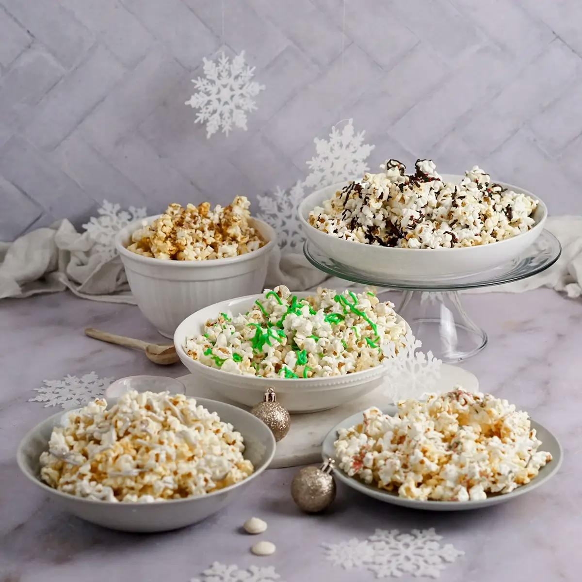 Five bowls of christmas popcorn drizzled with chocolate, peppermint, and other holiday toppings.