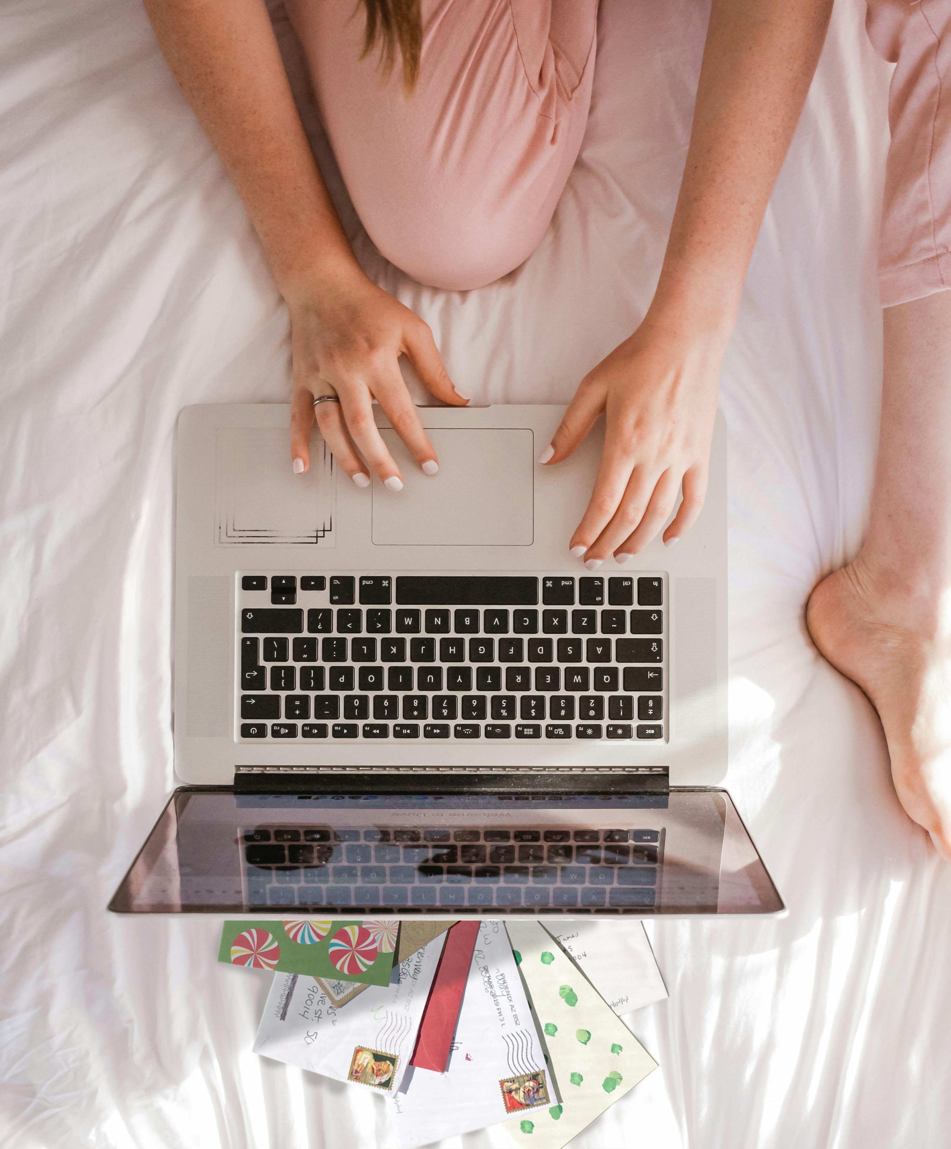 top down view of woman browsing her laptop on her bed with postcards laid out