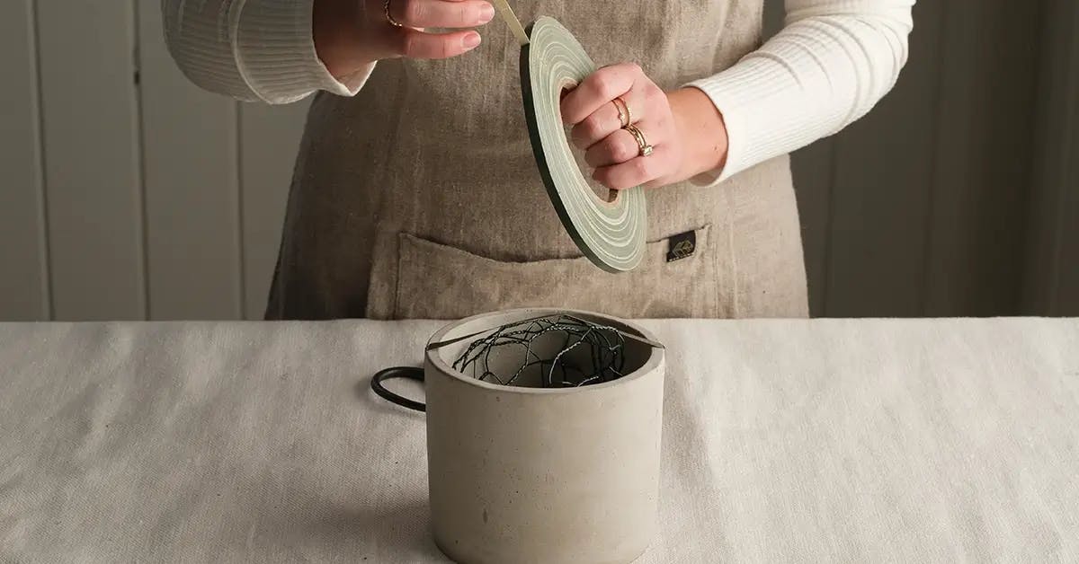 A vase filled with chicken wire, and hands unwrapping tape from a roll ready to secure the wire.