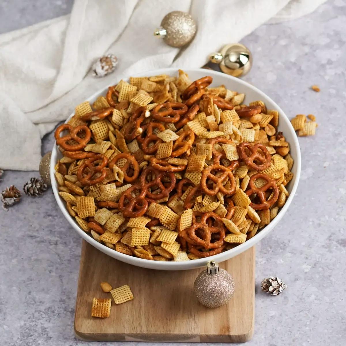 A bowl of savory gluten-free chex mix on a cutting board, surrounded by Christmas ornaments.