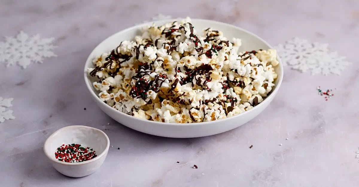 A bowl of Christmas popcorn drizzled with peppermint dark chocolate and topped with sprinkles.