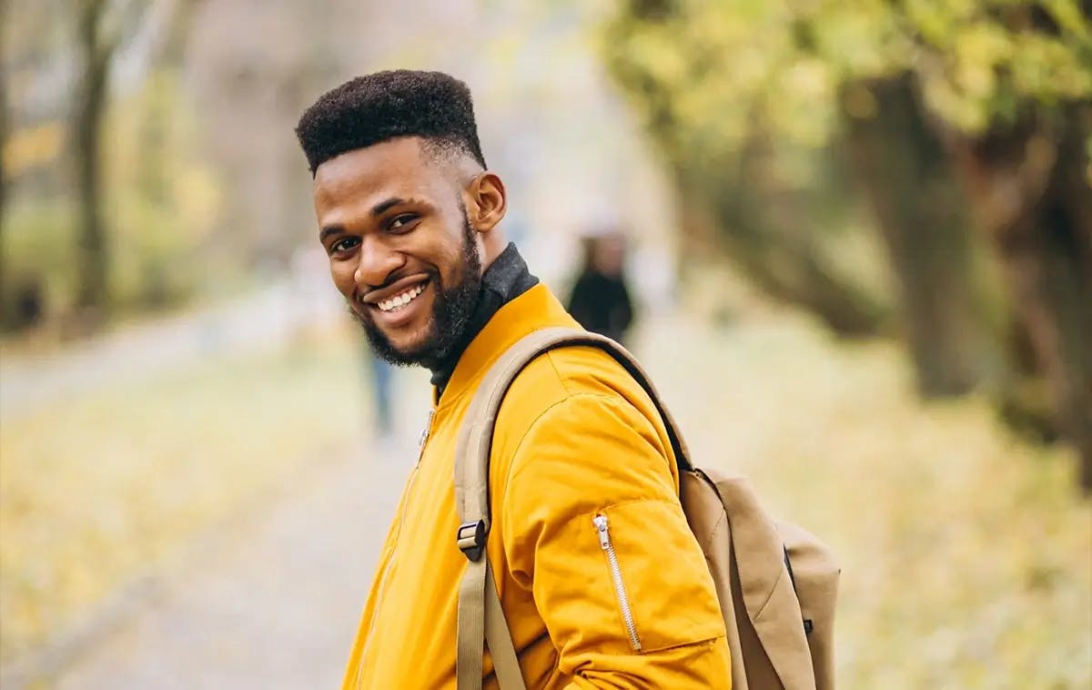 Black man in yellow jacket and backpack, standing in park and smiling at camera
