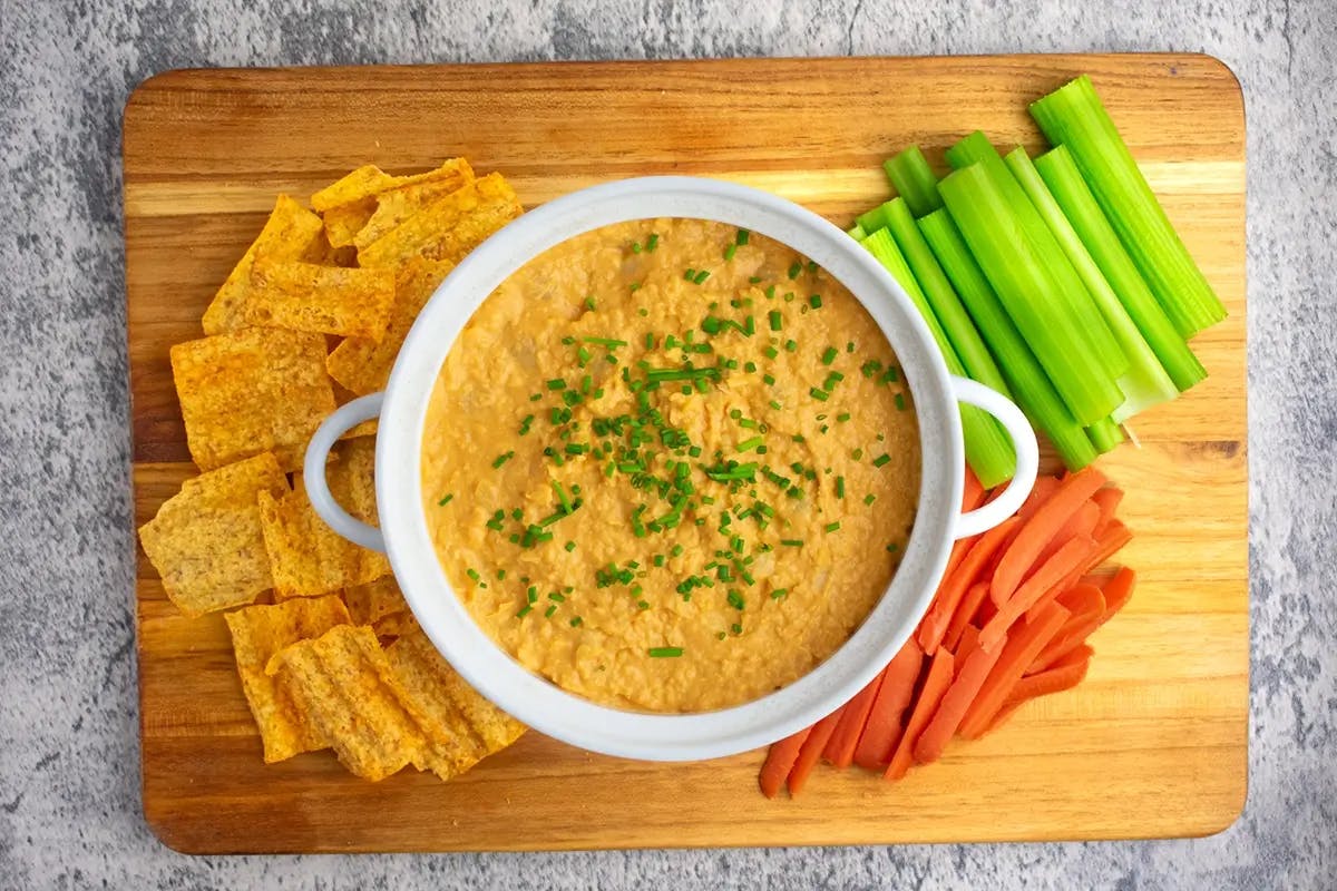 Bowl of vegan appetizer Buffalo Chickpea Dip on a wooden board, surrounded by celery, carrots and crackers.