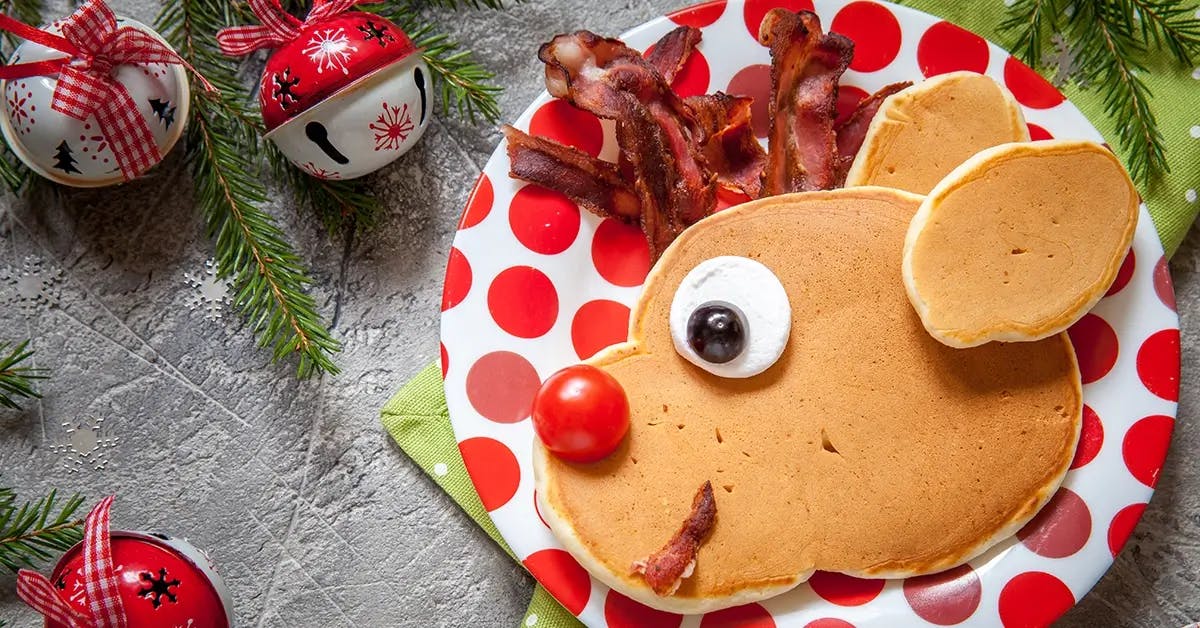 A Christmas breakfast pancake shaped to look like a reindeer, on a red & white spotted plate.
