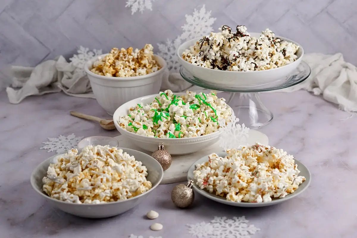 Five bowls of christmas popcorn drizzled with chocolate, peppermint, and other holiday toppings.