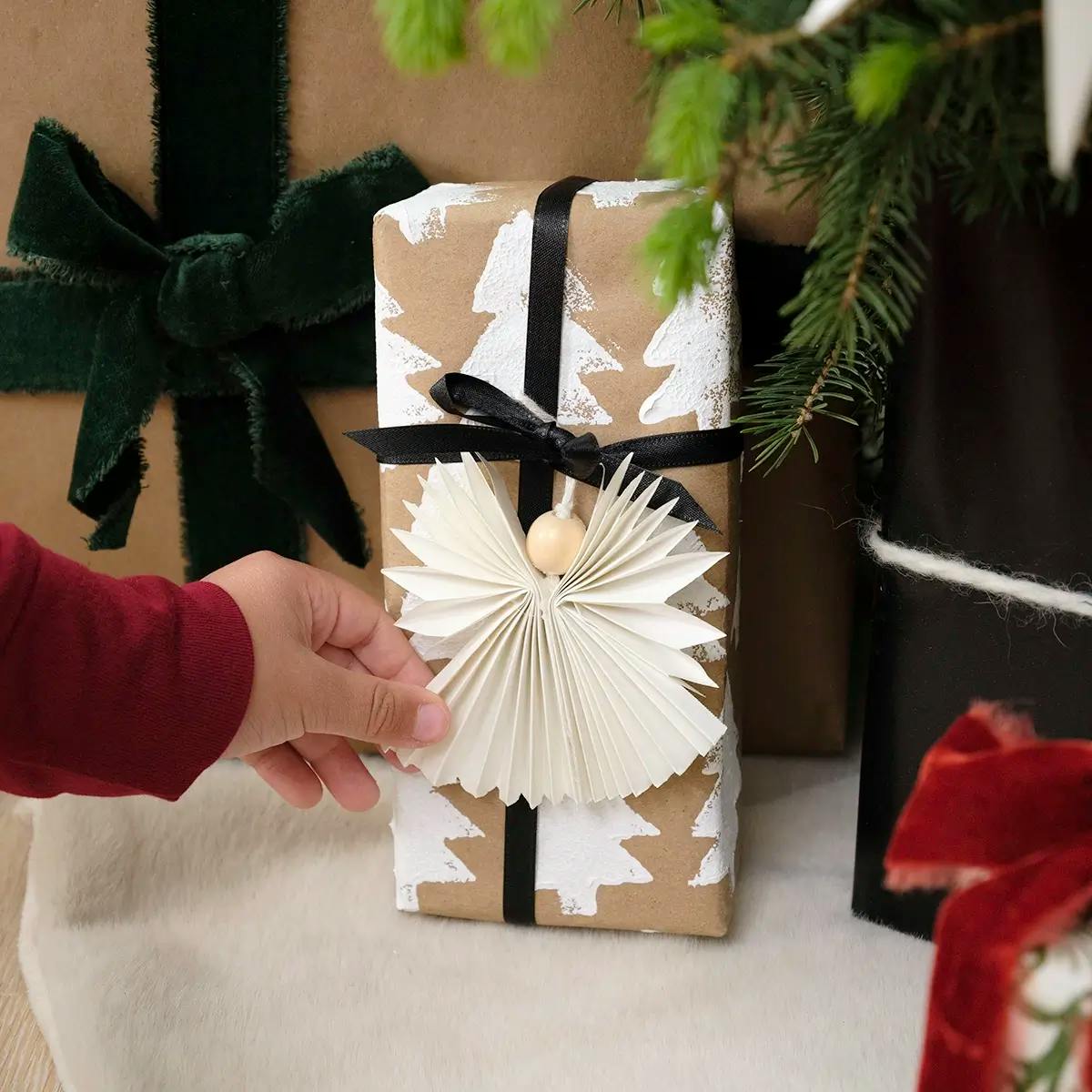 A hand pacing a gift under a Christmas tree. The gift is wrapped in custom Christmas wrapping paper made with e DIY stamp, and decorated with an angel ornament.