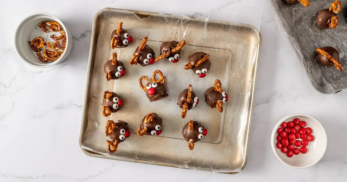 Reindeer Cookie Dough Bites on a baking sheet, covered in chocolate with pretzel pieces for antlers and mini M&Ms as eyes and nose.