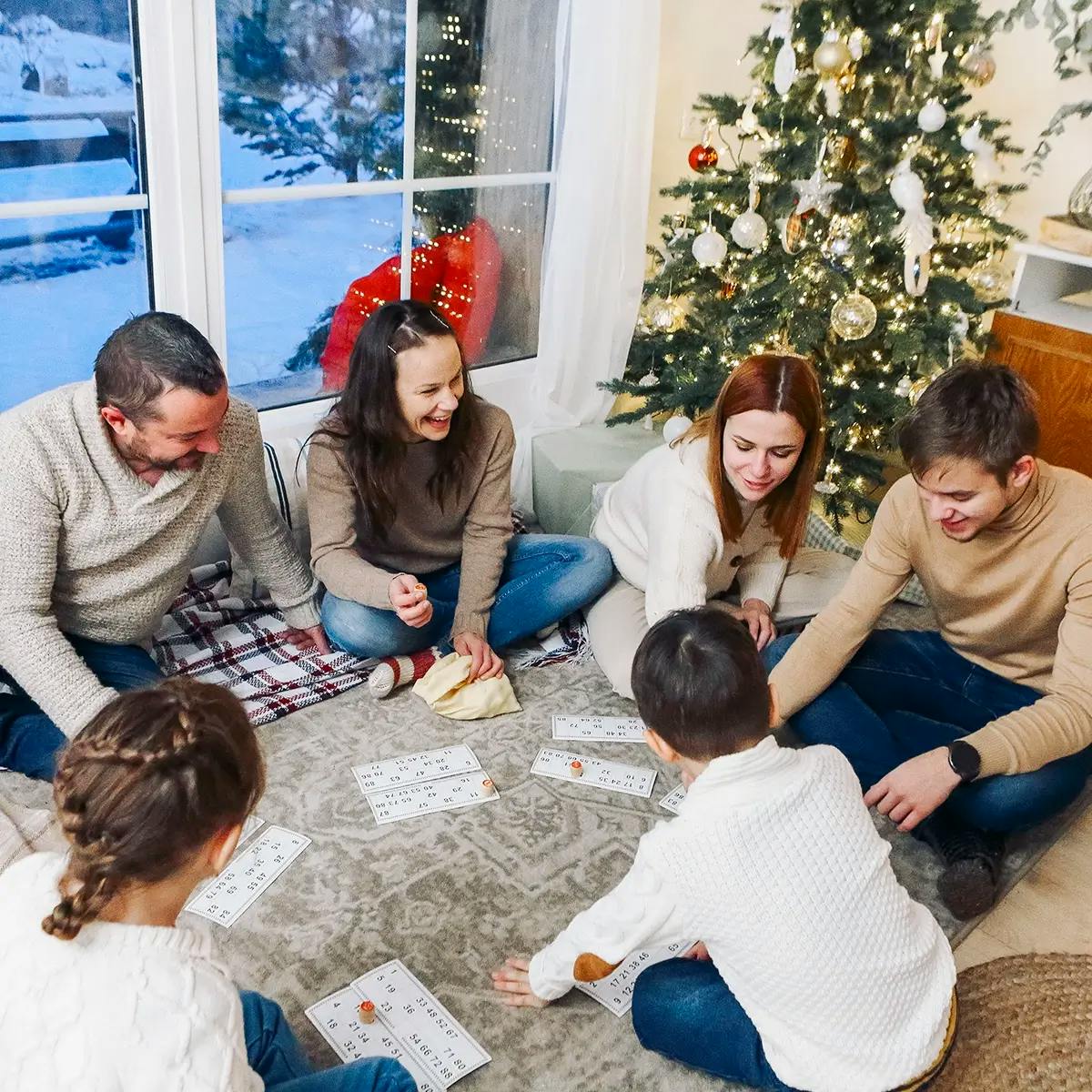 Adults and children sitting on the floor playing a bingo-like Christmas game, with snow outside the window and a Christmas tree in the background.