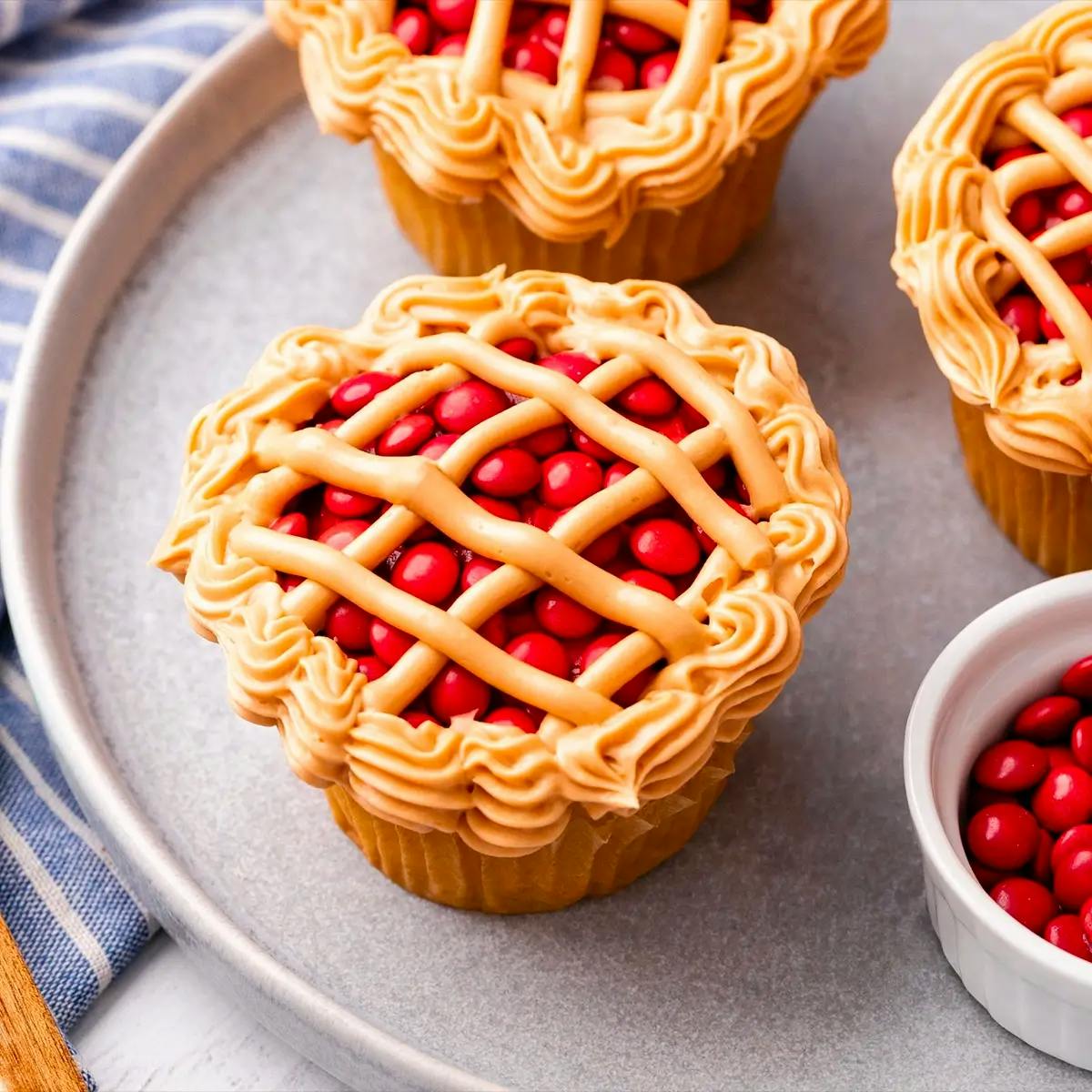 Cupcake topped with red M&Ms and piped with a lattice frosted crust that looks just like a mini cherry pie.