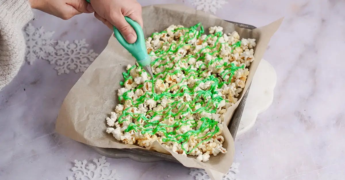 A hand drizzling green melted almond bark over a bowl of Christmas popcorn.