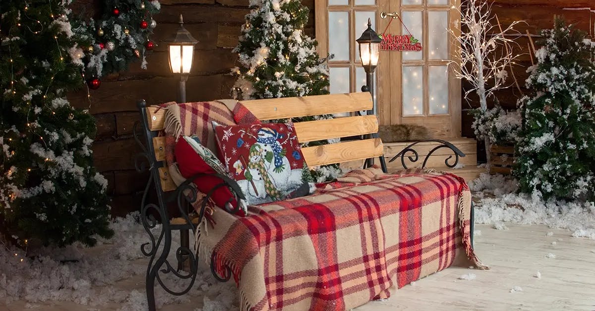 A rustic and festive front porch with a bench in the foreground, covered in a plaid blanket with christmas pillows.