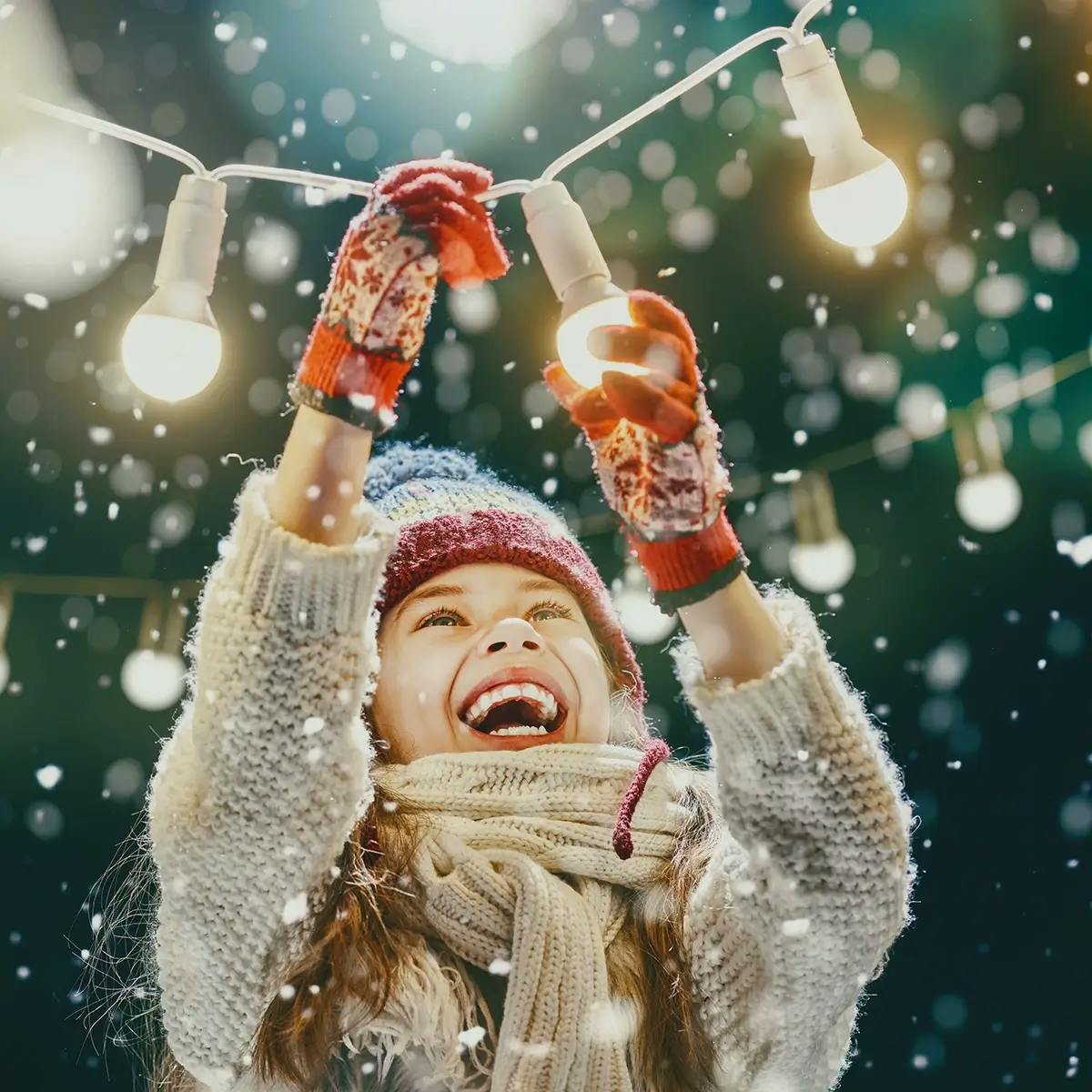 Woman outdoors in snow hanging lights, wearing a wool hat, sweater, and mittens.