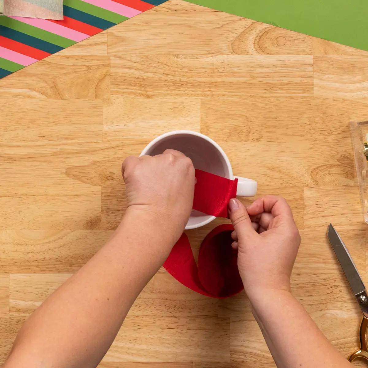Attaching the first strip of crepe paper to the mug with double-sided tape, as part of a tutorial on how to wrap a mug.