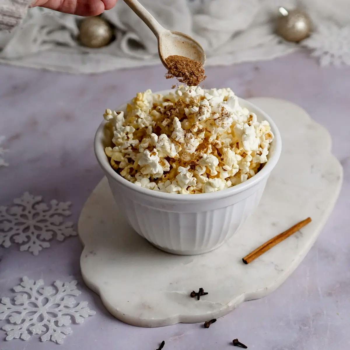 A hand spooning gingerbread spices over the top of a bowl of Christmas popcorn.