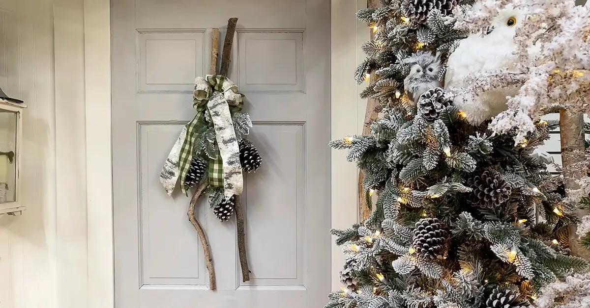 Front porch with white front door with a handmade wreath of branches and pinecones on the door. A frosted Christmas tree in foreground with acorns and bows.