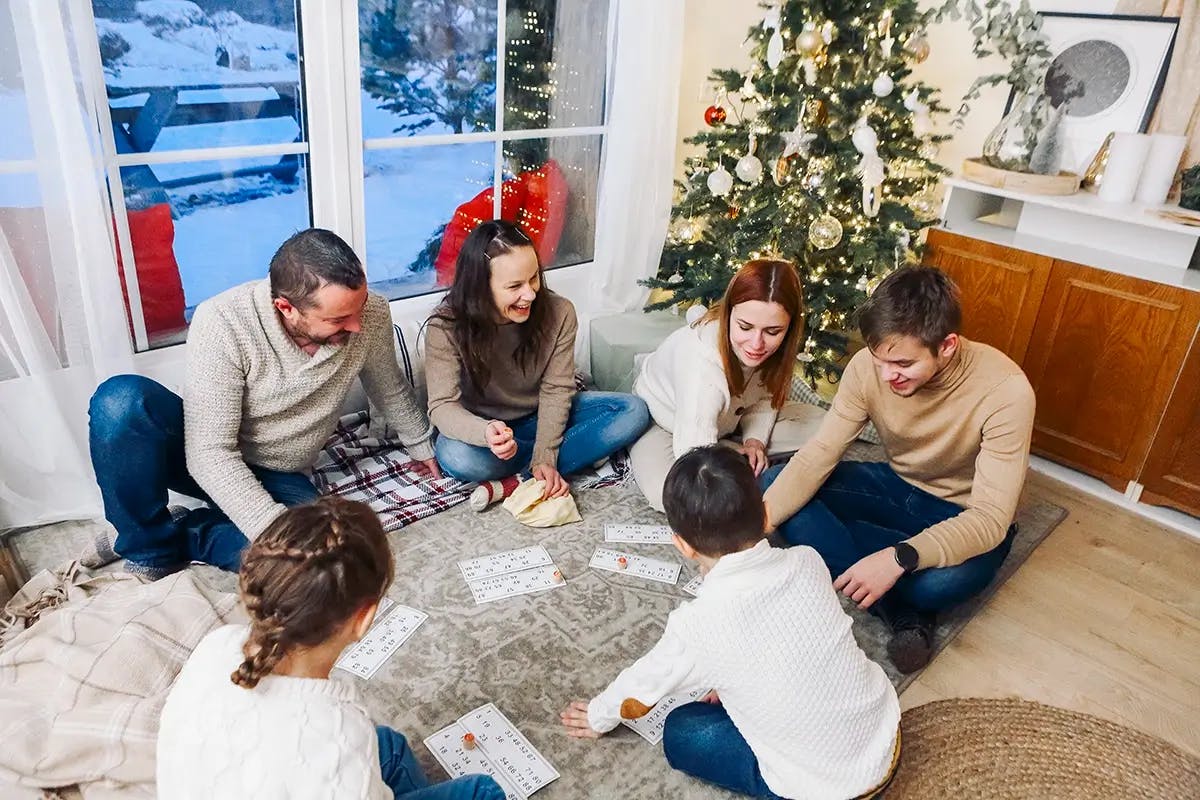 Adults and children sitting on the floor playing a bingo-like Christmas game, with snow outside the window and a Christmas tree in the background.