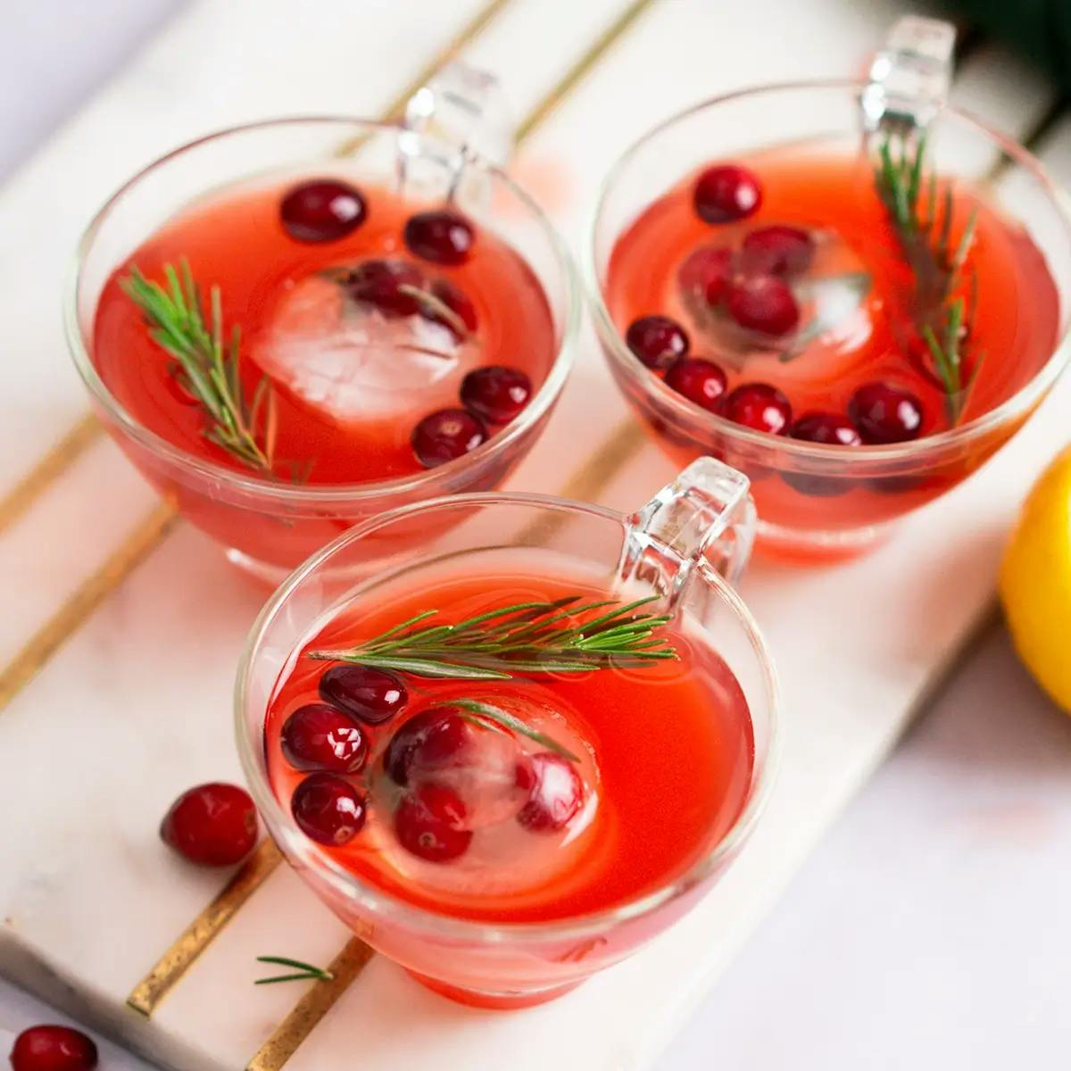 Three glasses of Christmas Punch on a cutting board next to half a lemon and a bowl of cranberries.