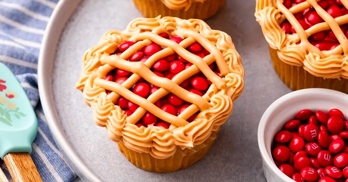 Cupcake topped with red M&Ms and piped with a lattice frosted crust that looks just like a mini cherry pie.