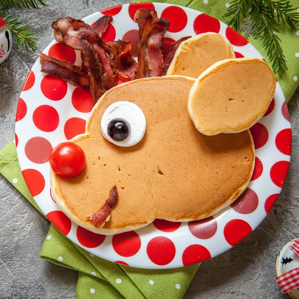 A pancake shaped like Rudolph on a red spotted plate, with bacon for antlers, mozzarella and an olive for eyes, bacon for a mouth and a cherry tomato for the nose.