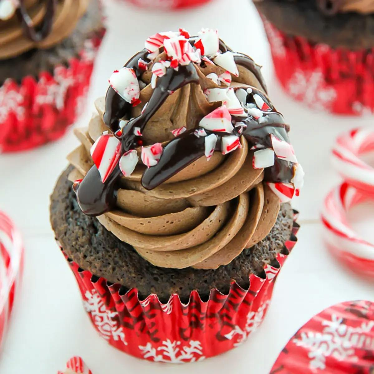 A chocolate cupcake covered in creamy light chocolate frosting, drizzled in dark chocolate sauce and sprinkled with crushed candy canes, with more blurred cupcakes in the background.