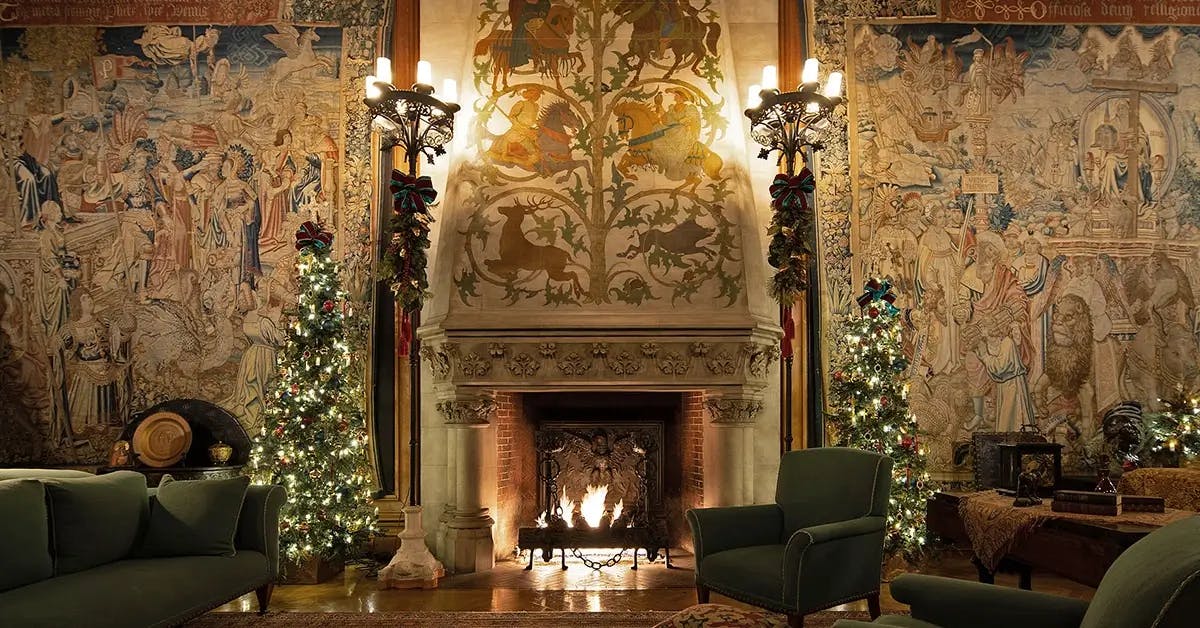 Grand room in the Biltmore Estate with fireplace flanked by two ornate tapestries, two Christmas trees, garland and lights.
