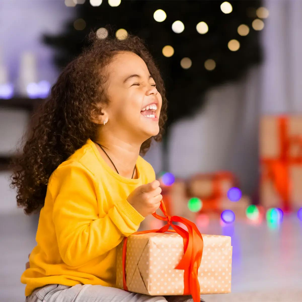 Excited Black girl laughing with Christmas gift, with blurred gifts and Christmas tree in background