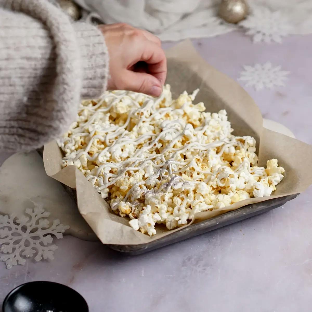 Christmas popcorn on a baking tray, drizzled with white chocolate. A hand is sprinkling edible silver glitter over the popcorn.