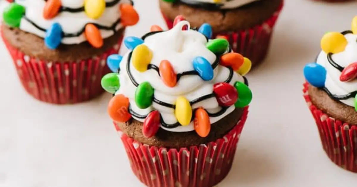 Chocolate cupcakes topped with white frosting, drizzled with brown decorative frosting to imitate wires, with multi-colored M&Ms as Christmas lights.