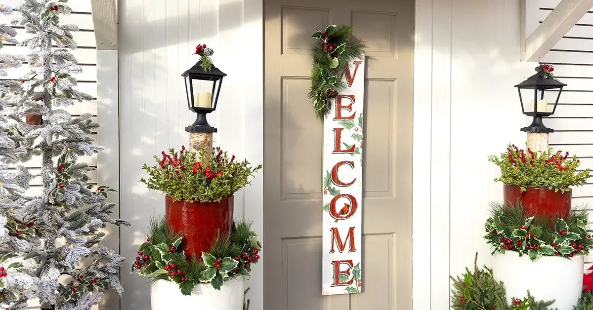 Festive front porch with large welcome sign on door, with festive Christmas pots, plants and garland surrounding door, porch, rails and windows.